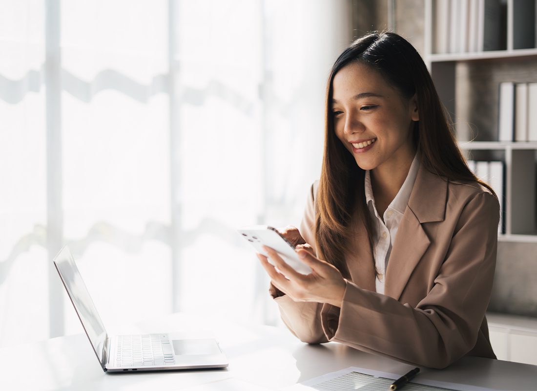 Blog - Professional Smiling at Her Phone While Sitting at Her Desk with Her Laptop Open and She is Typing on Her Mobile in Her Hand